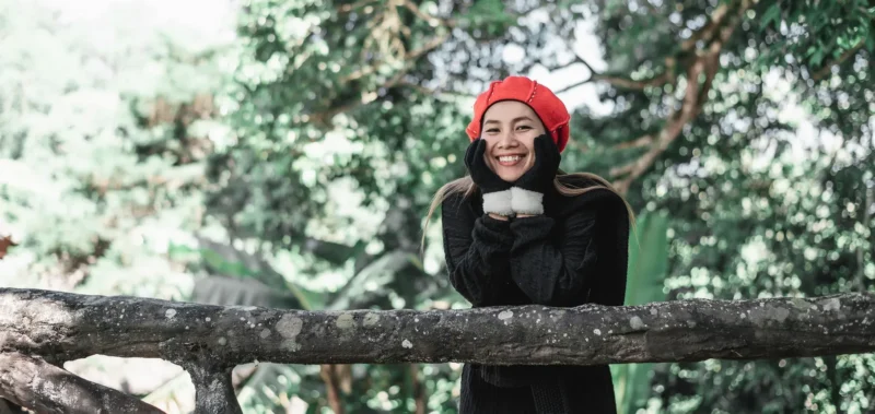 Femme joyeuse en béret rouge devant une balustrade dans un écrin naturel verdoyant.